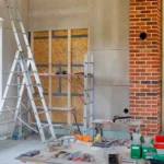 Eine Baustelle in einem Altbau, bei der Wände mit Trockenbau und Innendämmung verkleidet werden, davor stehen eine Leiter und Werkzeuge.