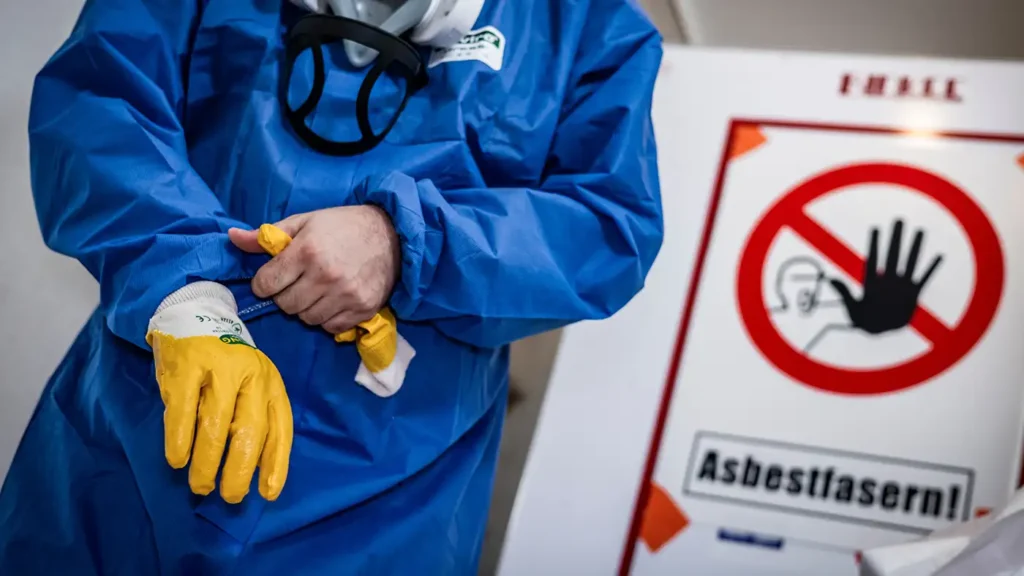 Eine Person im blauen Schutzanzug streift sich gelbe Sicherheitshandschuhe über; im Hintergrund steht ein weißes Warnschild mit Verbotszeichen und der Aufschrift „Asbestfasern!“.