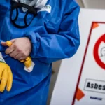 Eine Person im blauen Schutzanzug streift sich gelbe Sicherheitshandschuhe über; im Hintergrund steht ein weißes Warnschild mit Verbotszeichen und der Aufschrift „Asbestfasern!“.